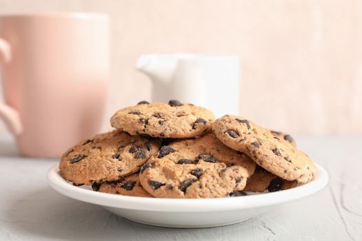 Plate with tasty chocolate chip cookies and blurred cup of milk on gray background, closeup