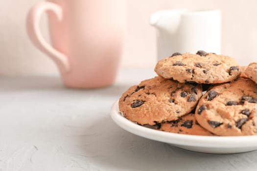 Plate with tasty chocolate chip cookies and blurred cup of milk on gray background, closeup
