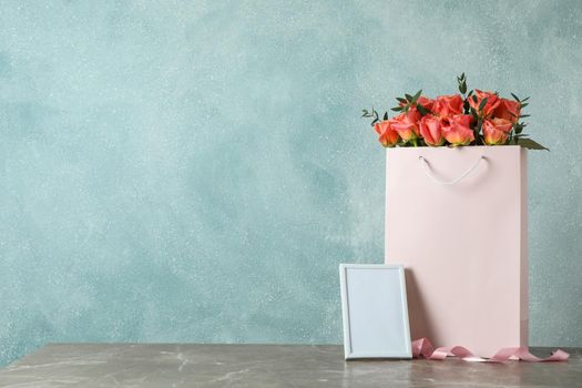 Gift bag with bouquet of pink roses and empty frame on grey table against light background