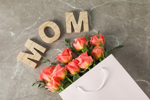 Gift bag with bouquet of orange roses and inscription Mom on brown background, top view