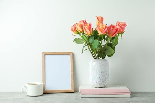Vase with pink roses, copybooks, empty frame, cup of coffee on grey table against white background