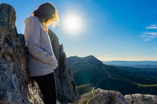 Lonely woman finds herself leaning against the rocks in the nature pensive at top of mountains with a splendid view below and sun through her hair. Concept of positive loneliness to find your own way