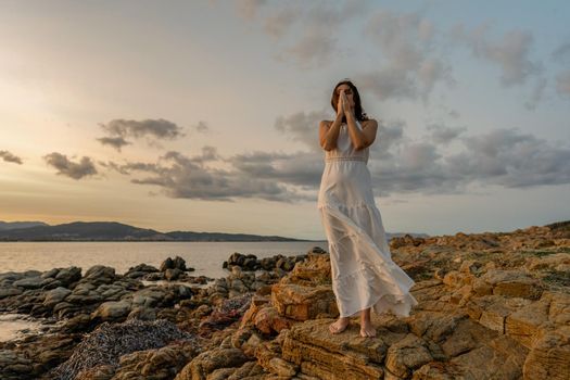 Pensive Woman in long white dress on with folded hands in front of face and closed eyes contemplating the sunset over the sea. Concept of experiencing nature to feel good and find your spirituality