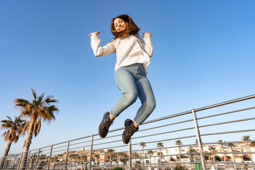 Happy smiling girl jumping outdoor high with arms up looking down at the camera with her hair ruffled by the wind. Concept of happiness and good life emotion for success of positive people in youth