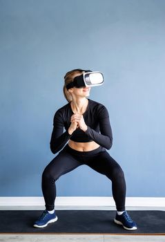 Fitness, sport and technology. Young athletic woman wearing virtual reality glasses squatting at blue background