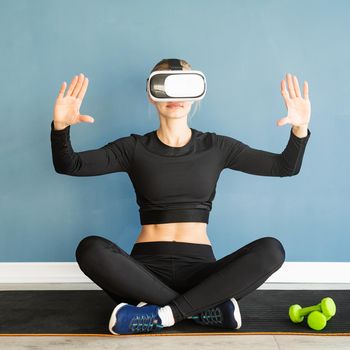 Fitness, sport and technology. Young athletic woman wearing virtual reality glasses sitting at fitness mat using VR interactive menu at blue background