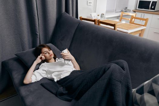 a woman under a warm blanket lies on a sofa and a window in the background. High quality photo