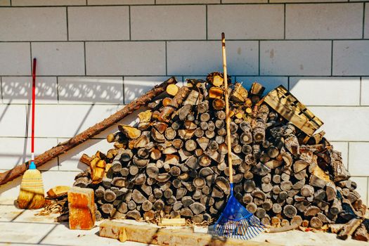 A mountain of chopped wood for the fireplace near the wall. Broom and rake near a mountain of firewood.