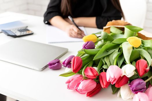 A young business woman florist writing in notebook or calendar at the office, bucket of tulips on the desk. Small business.