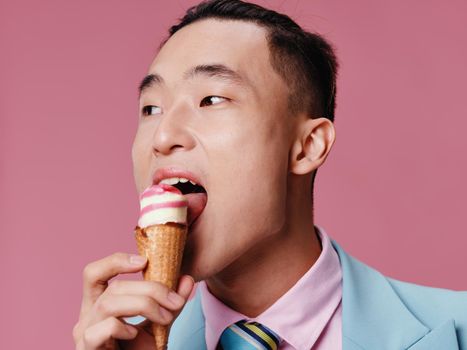 Happy Asian man eating ice cream on pink background in blue classic suit cropped view. High quality photo
