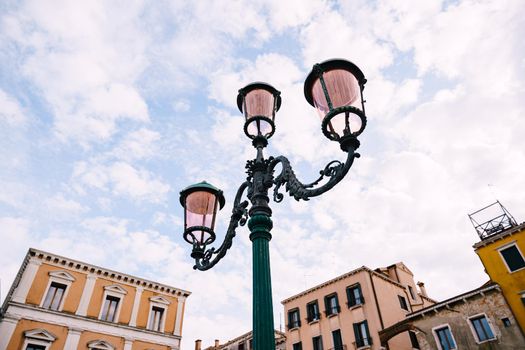 An ancient triple metal street lamp, with pink plafonds against the sky of Venice, Italy. Vintage street lighting, elegant pillar design.