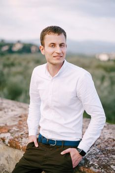 A large male portrait on the roof of an old winery villa in Tuscany, Italy. White long-sleeved shirt, smart watch, blue belt, hands in pants pockets.