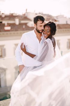 Mixed-race wedding couple. Destination fine-art wedding in Florence, Italy. African-American bride hugs Caucasian groom on the roof with cityscape view.