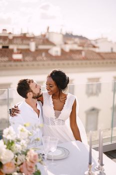 Interracial wedding couple. Destination fine-art wedding in Florence, Italy. African-American bride and Caucasian groom are sitting at rooftop wedding dinner table overlooking the city.