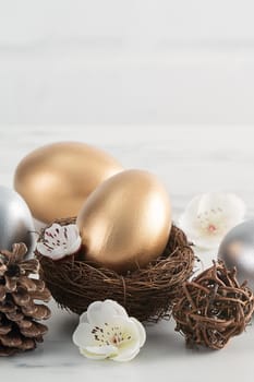 Close up of golden and silver Easter eggs in the nest with white plum flower on bright white wooden table background.