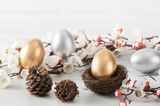 Close up of golden and silver Easter eggs in the nest with white plum flower on bright white wooden table background.