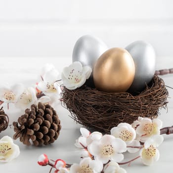 Close up of golden and silver Easter eggs in the nest with white plum flower on bright white wooden table background.