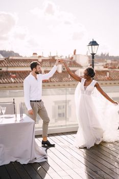 Interracial wedding couple. Destination fine-art wedding in Florence, Italy. African-American bride and Caucasian groom dance near the table for a wedding dinner, on the roof of the restaurant.