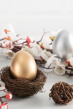 Close up of golden and silver Easter eggs in the nest with white plum flower on bright white wooden table background.