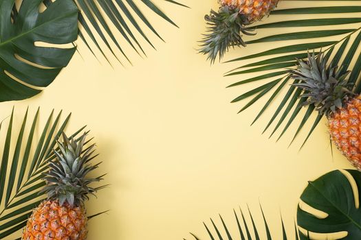 Top view of fresh pineapple with tropical palm and monstera leaves on yellow table background.