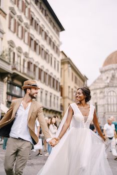African-American bride and Caucasian groom run along Piazza del Duomo. Multiracial wedding couple. Wedding in Florence, Italy.