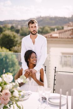 Interracial wedding couple. Destination fine-art wedding in Florence, Italy. African-American bride sits at wedding table, Caucasian groom hugs her shoulders.