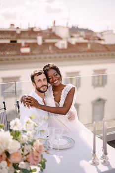 Interracial wedding couple. Destination fine-art wedding in Florence, Italy. African-American bride and Caucasian groom are sitting at rooftop wedding dinner table overlooking the city.