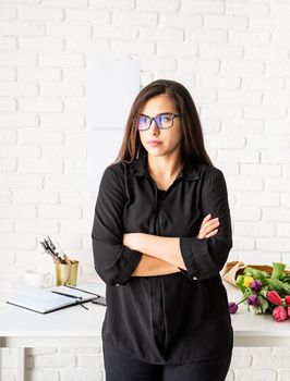 Small business concept. Portrait of young confident brunette business woman working at the office, standing by the desk with arms crossed