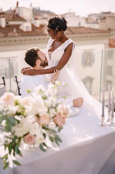 Interracial wedding couple. Destination fine-art wedding in Florence, Italy. roof, table, banquet, dinner, An African-American bride sits on her lap at a Caucasian wedding table on the roof.