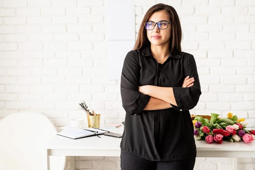 Small business concept. Portrait of young confident brunette business woman working at the office, standing by the desk with arms crossed