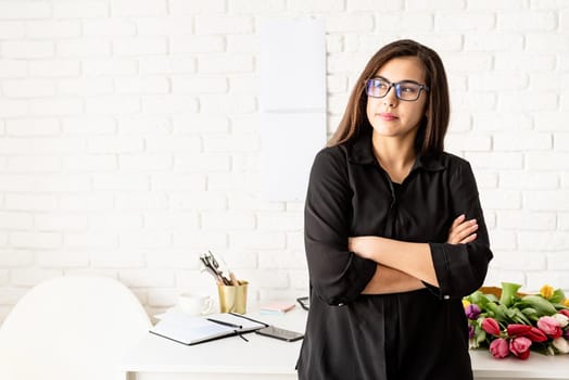 Small business concept. Portrait of young confident brunette business woman working at the office, standing by the desk with arms crossed