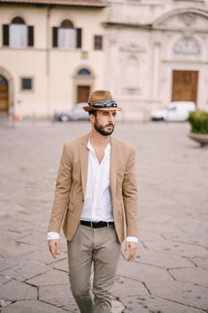 A young guy with a straw hat, a white unbuttoned shirt and a sand jacket, a small beard walks around of Florence.