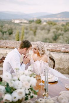 The wedding couple sits at the dinner table on the roof of the old villa, the groom holds the bride's hands. Wedding at an old winery villa in Tuscany, Italy