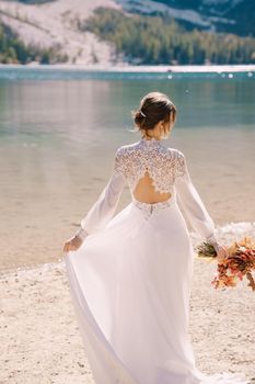 Beautiful bride in a white dress with sleeves and lace, with a yellow autumn bouquet of dried flowers and peony roses, at Lago di Braies in Italy. Destination wedding in Europe, at Braies lake.