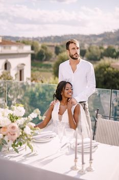 Mixed-race wedding couple. Destination fine-art wedding in Florence, Italy. African-American bride sits at wedding table, Caucasian groom hugs her shoulders.