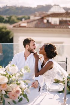 Interracial wedding couple. Destination fine-art wedding in Florence, Italy. African-American bride and Caucasian groom are sitting at rooftop wedding dinner table overlooking the city.