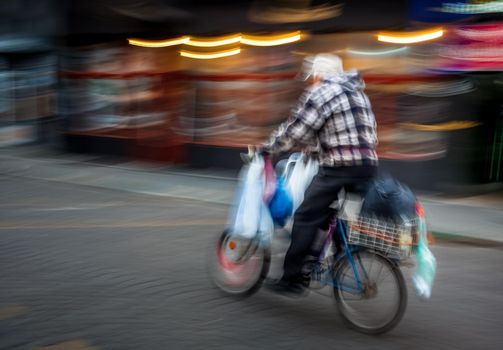 Old man with a large number of packets riding a bicycle around the city. Intentional motion blur