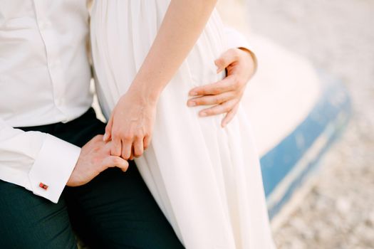 Bride and groom sit embracing and holding hands on an inverted boat, close-up. High quality photo