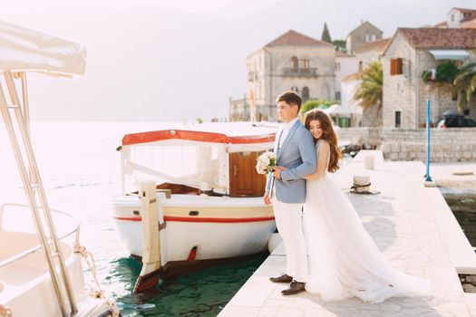 The bride hugs the groom from behind on the pier near the old town of Perast, next to them is a tourist boat . High quality photo