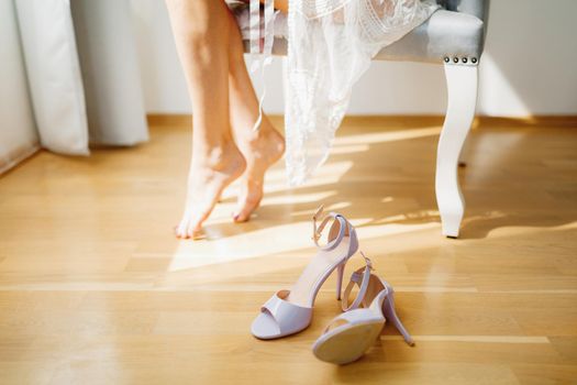 Barefoot bride sits in a chair in a hotel room during wedding preparations, sophisticated sandals are on the floor . High quality photo