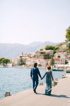 The bride in a stylish blue dress and groom walk along the pier holding hands near the old town of Perast, back view . High quality photo