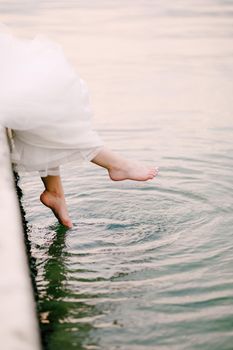 The bride sits on the pier and dangles her bare feet in the water, the legs peek out from under the wedding dress . High quality photo