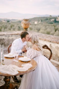 The wedding couple sits at the dinner table, on the roof of the villa and kisses. Wedding at an old winery villa in Tuscany, Italy.