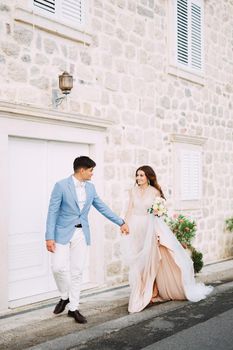 The bride and groom stand holding hands in front of a beautiful white house in the old town of Perast . High quality photo