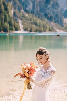 Beautiful bride in a white dress with sleeves and lace, with a yellow autumn bouquet of dried flowers and peony roses, at Lago di Braies in Italy. Destination wedding in Europe, at Braies lake.