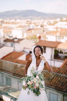 An African-American bride in a white dress, stands on the roof terrace, with a bouquet in her hands and in short gloves. Fine-art wedding in Florence, Italy
