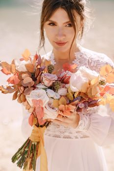 Beautiful bride in a white dress with sleeves and lace, with a yellow autumn bouquet of dried flowers and peony roses, at Lago di Braies in Italy. Destination wedding in Europe, at Braies lake.