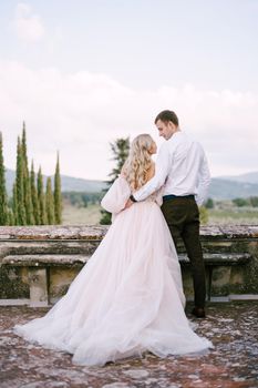 Wedding at an old winery villa in Tuscany, Italy. The wedding couple stands on the roof of an old winery, cuddling, standing with their backs in the frame.