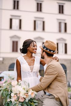 African-American bride in a white dress with a long veil and bouquet, and Caucasian groom in a sand jacket and straw hat. Interracial wedding couple. Wedding in Florence, Italy.