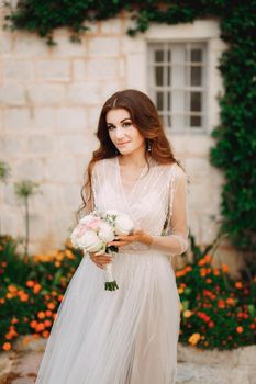 A bride with a bouquet in her hands stands at the wall of a house with a green liana and orange flowers in Perast . High quality photo